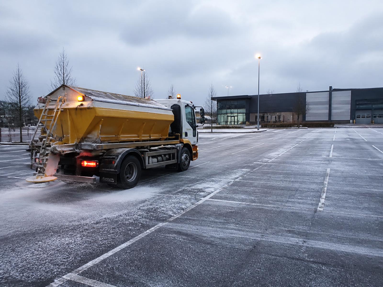 Commercial gritting vehicle treating a frosted retail park car park at dawn